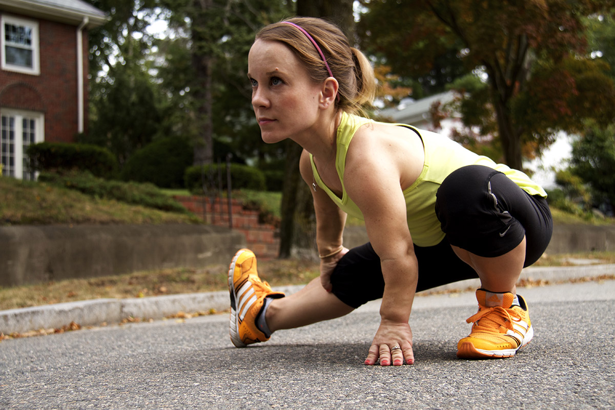 A woman with dwarfism, a condition that results in short stature, is dressed in athletic clothes. She is stretching on a residential street, preparing for a run
