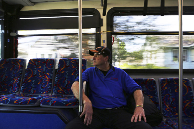 A man in a baseball hat and blue shirt rides a city bus alone, with trees and houses rushing by the windows