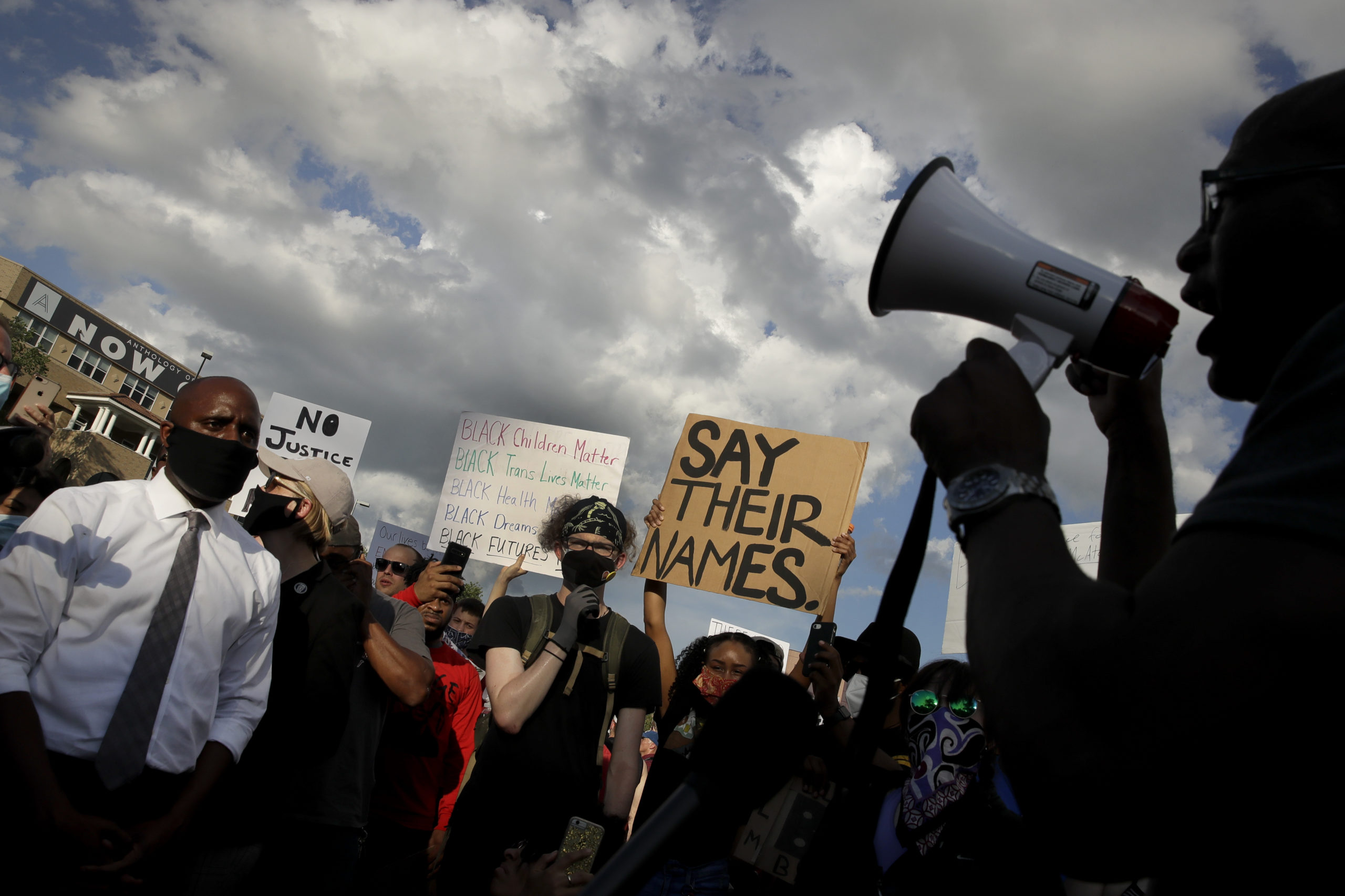 A crowd of people hold up signs One reads "Say Their Names," another reads "Black Children Matter. Black Trans Lives Matter. Black Health Matters. Black Dreams Matter. Black Futures Matter." A third sign, partially blocked by the crowd, reads "No Justice." To the right of the frame, Mayor Lucas yells into a megaphone.