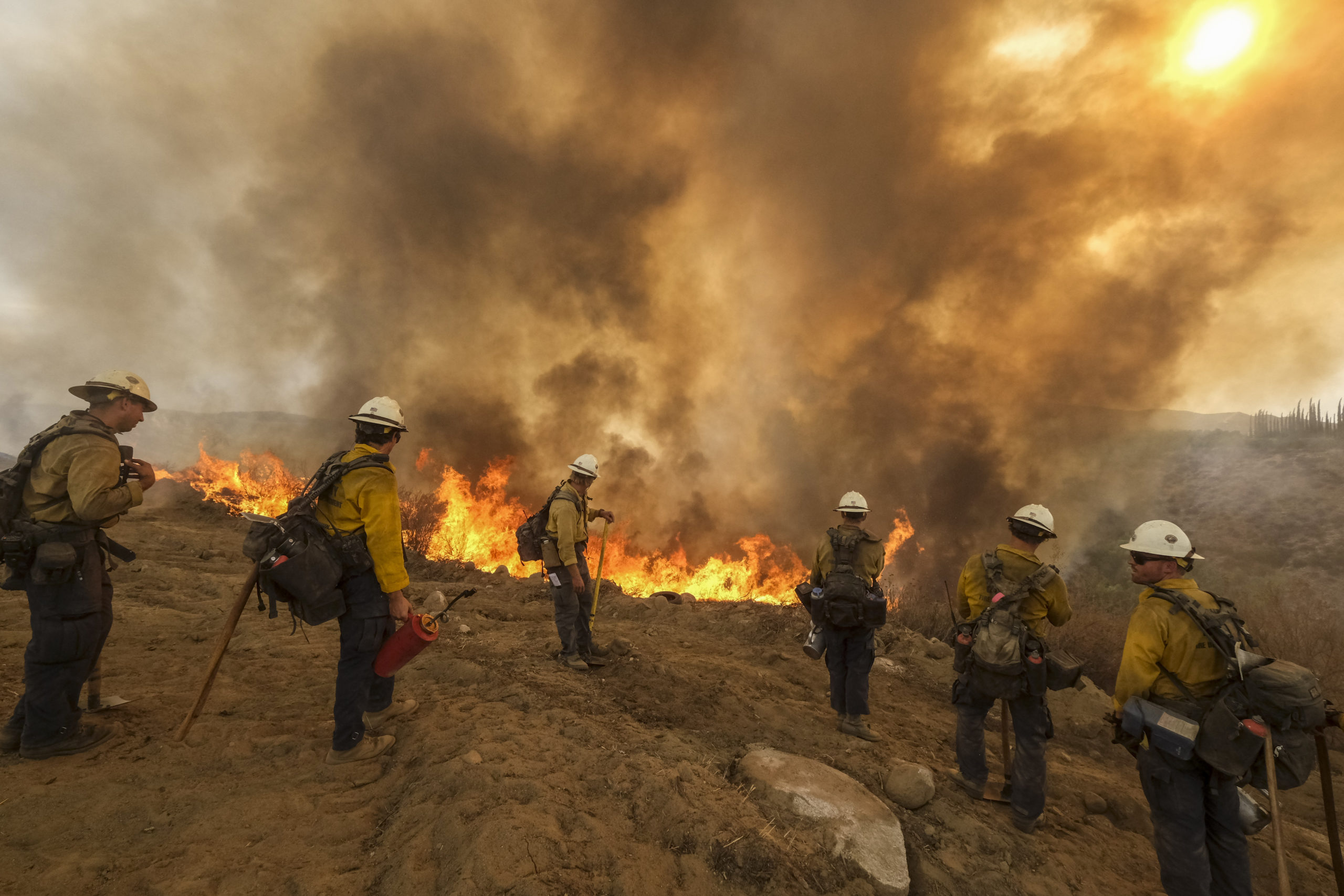 Four firefighters stand at the top of a hill overlooking a wildfire. A large cloud of black smoke hovers over them.
